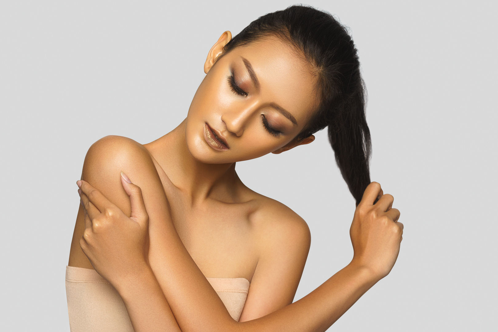 Beauty portrait with golden-toned skin and pulled-back hair, eyes closed, against a gray background