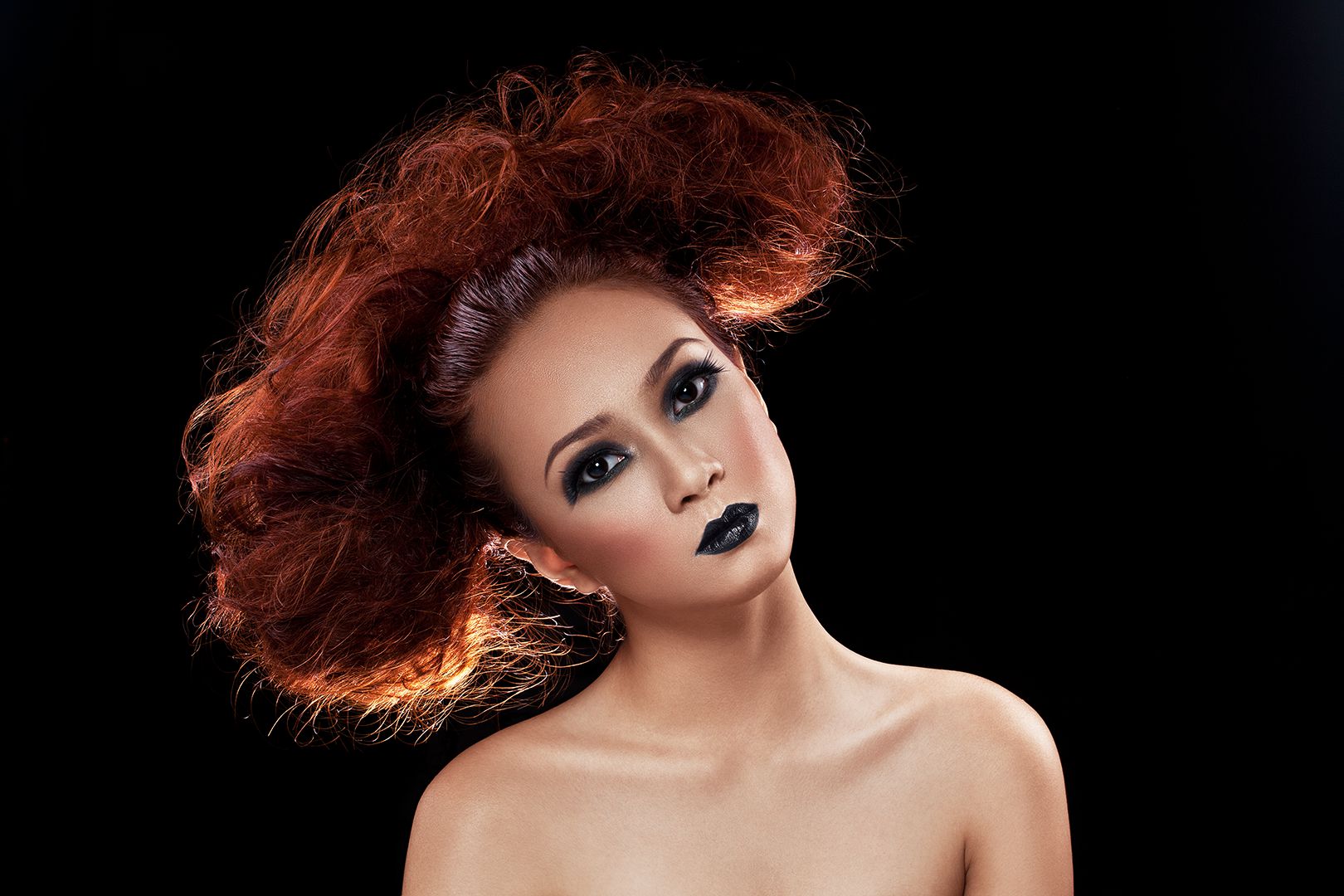 Beauty portrait with voluminous red hair, smoky black eye makeup, and black lipstick against black background