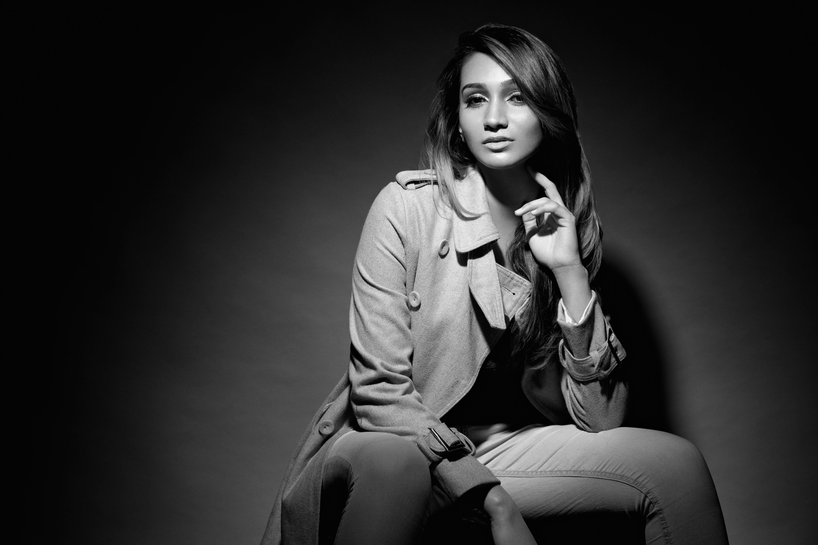 Black and white fashion portrait seated in trench coat with dramatic studio lighting