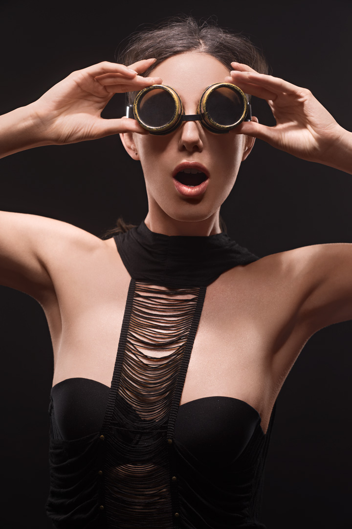 Editorial fashion portrait with brass steampunk goggles and shredded black bodysuit against dark background