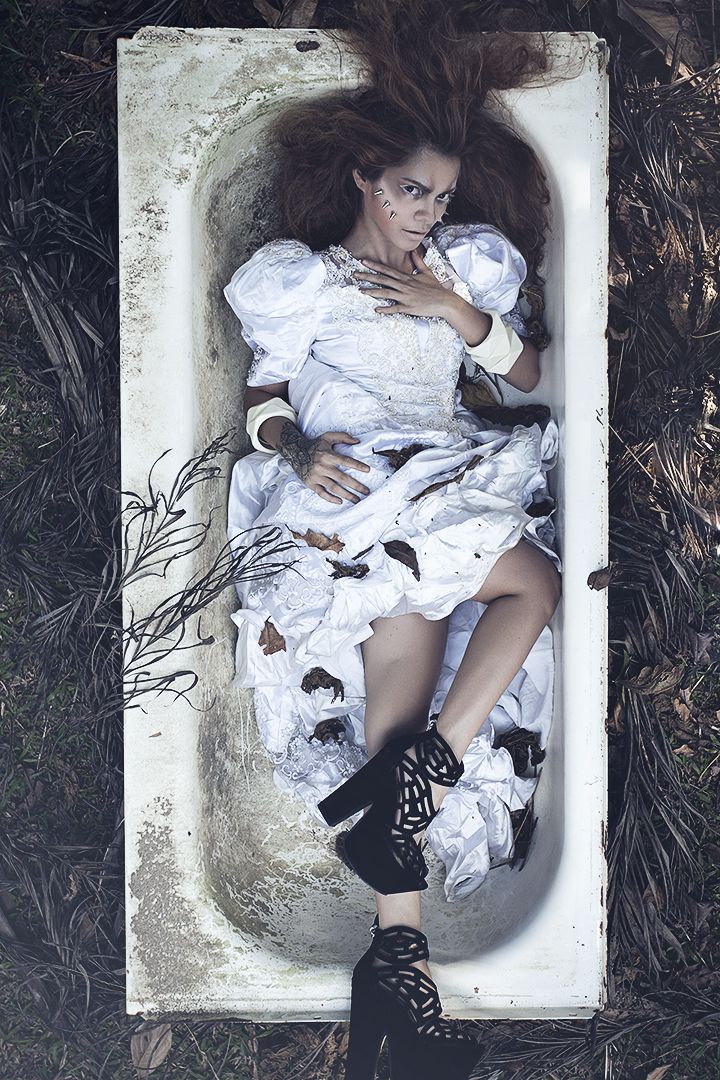 Conceptual editorial shot from overhead of model in deconstructed white dress lying in abandoned bathtub