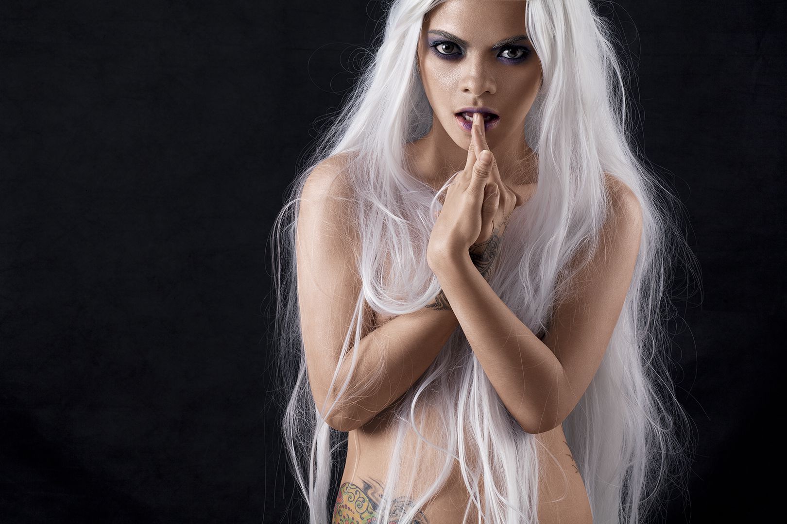 Moody intimate portrait with platinum white hair, tattoos, and dark smoky eye makeup against black